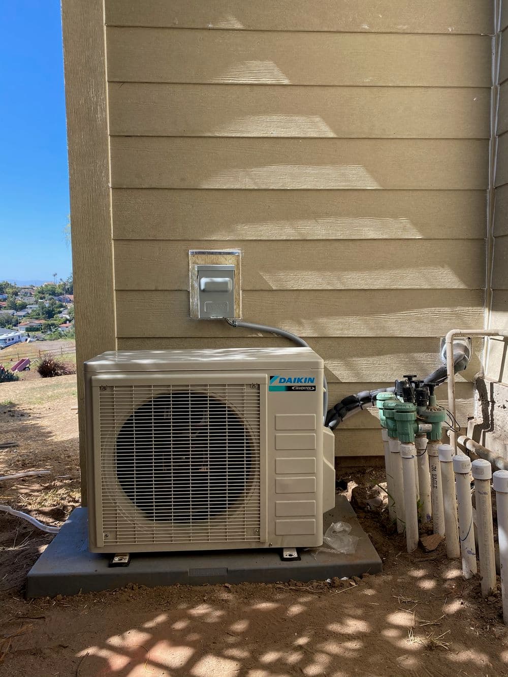 Daikin air conditioning unit installed outside a building, with exposed piping and utility outlet.