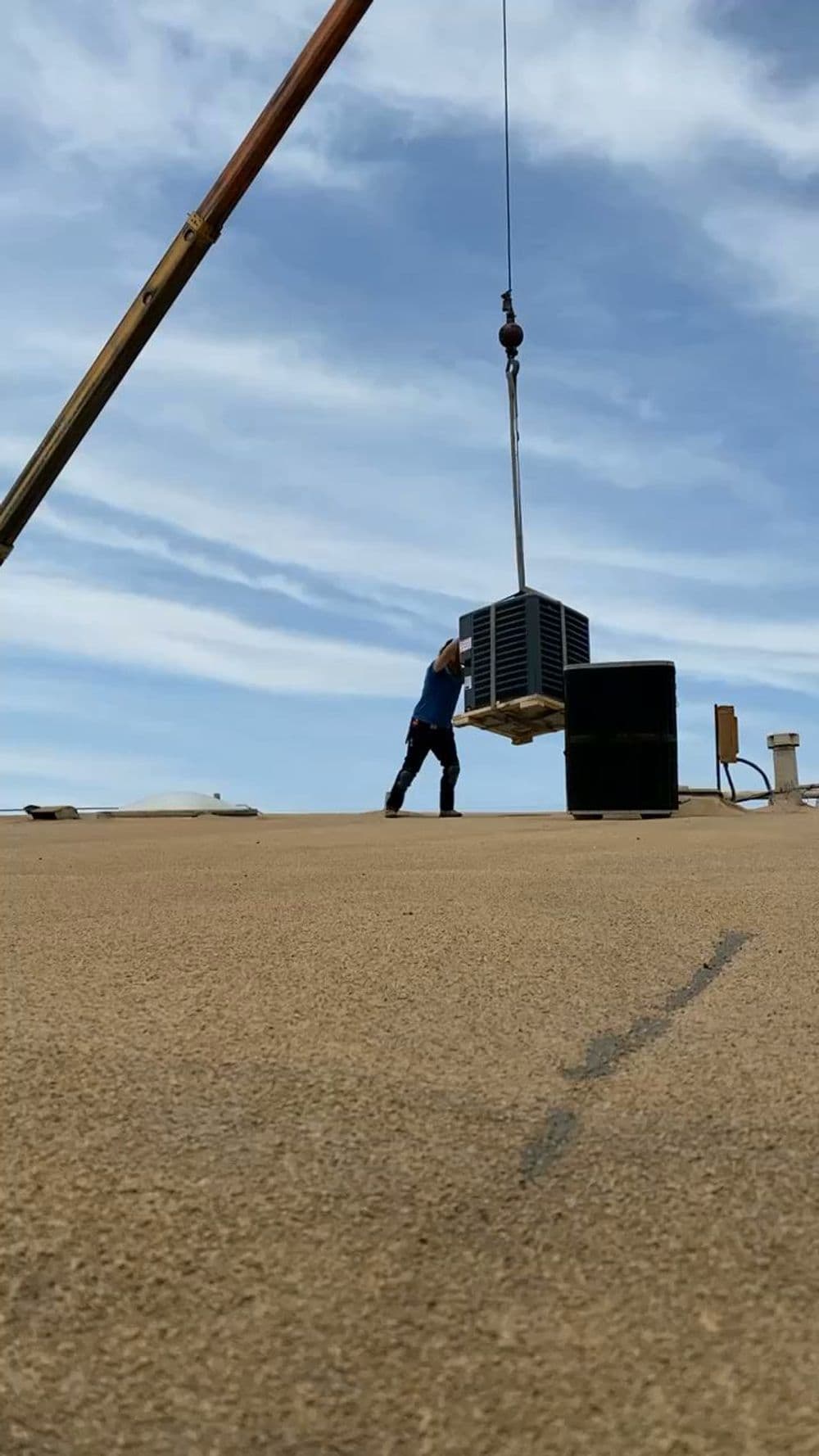 Technician using crane to install HVAC unit on rooftop under blue sky.