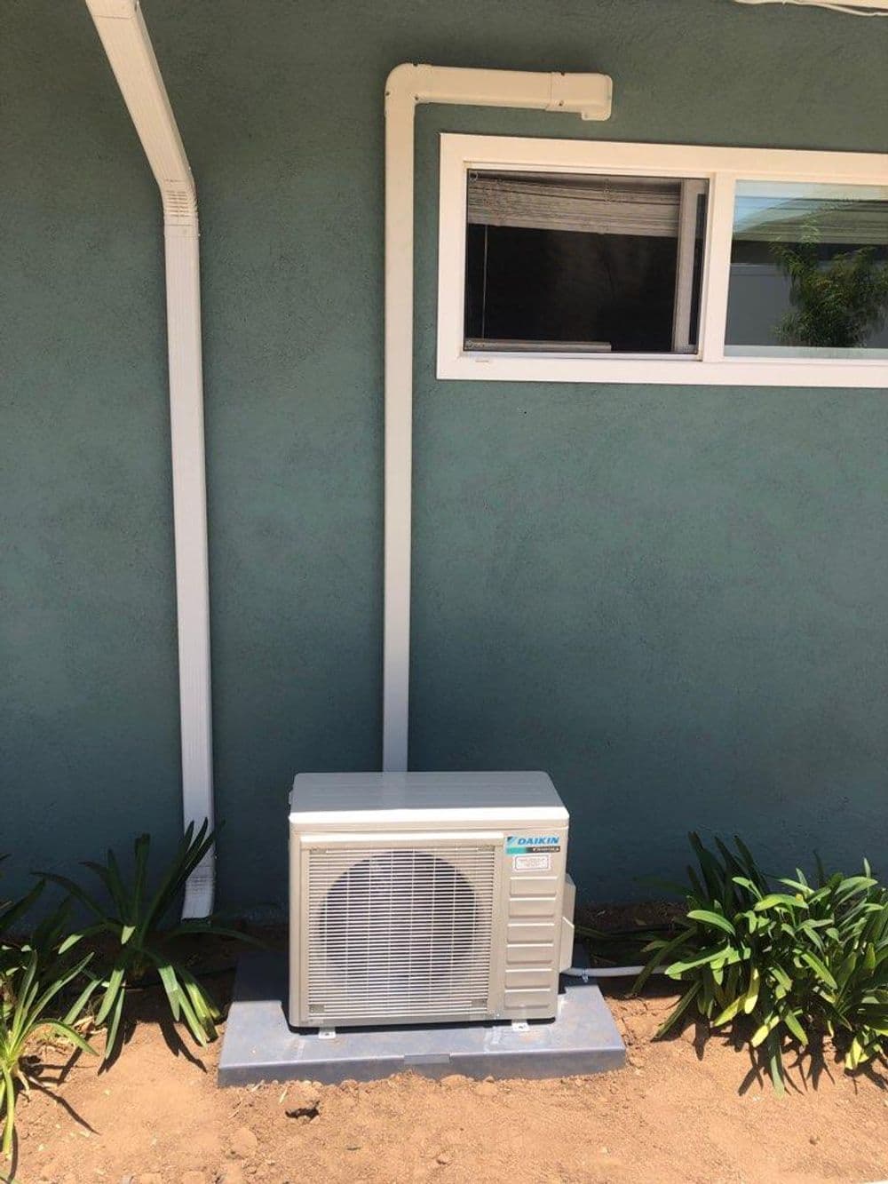 Air conditioning unit installed against a green wall, featuring nearby plants and drainage pipes.