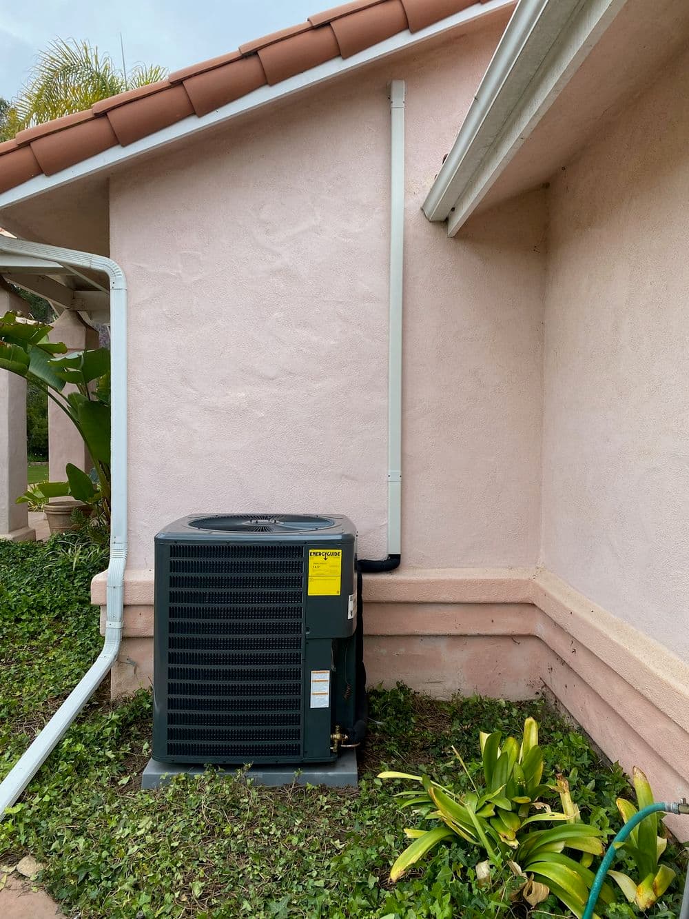 Air conditioning unit installed on a residential wall surrounded by greenery and pink stucco.