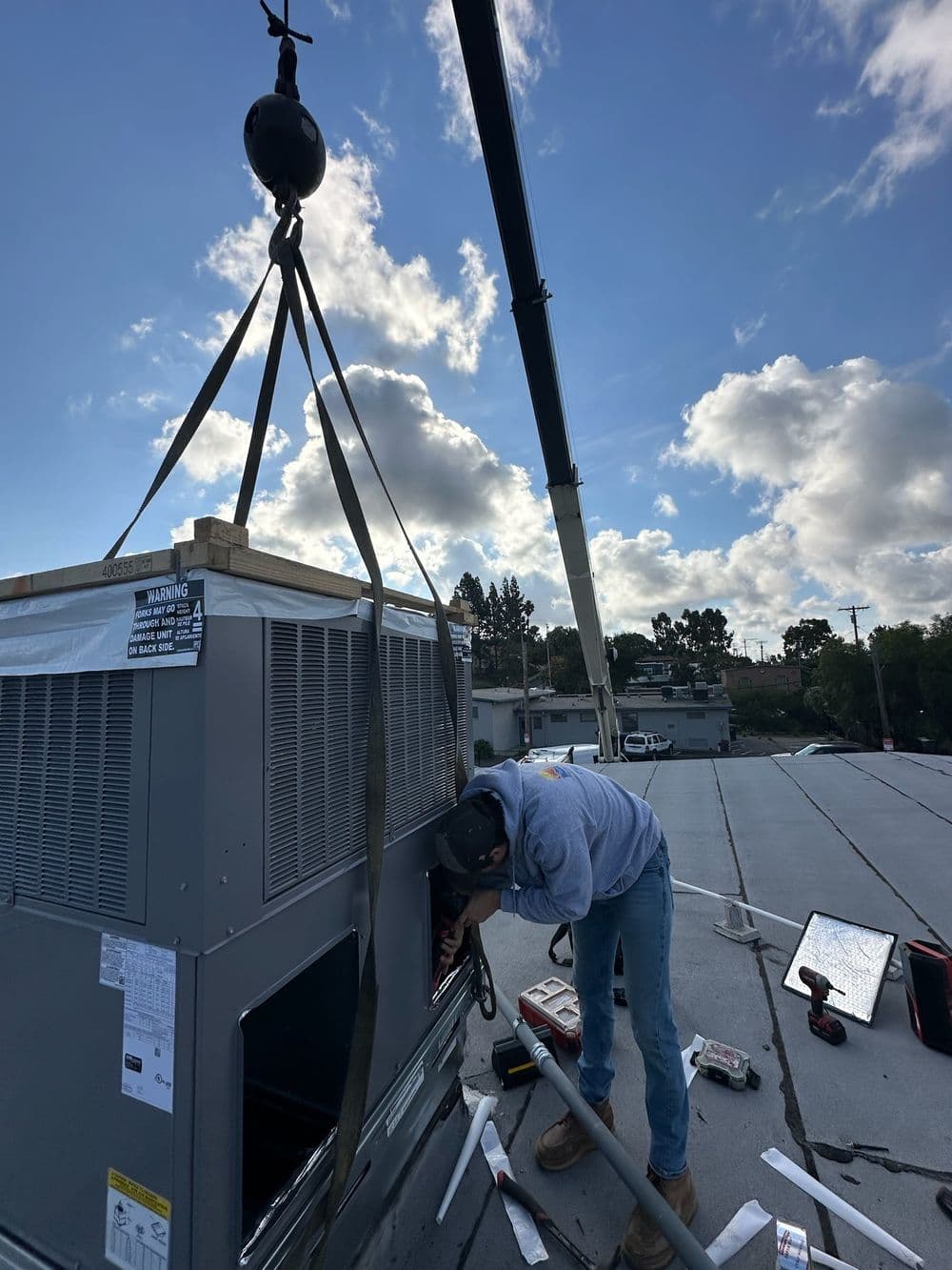 Technician installing HVAC unit on rooftop with crane against a blue sky and clouds.