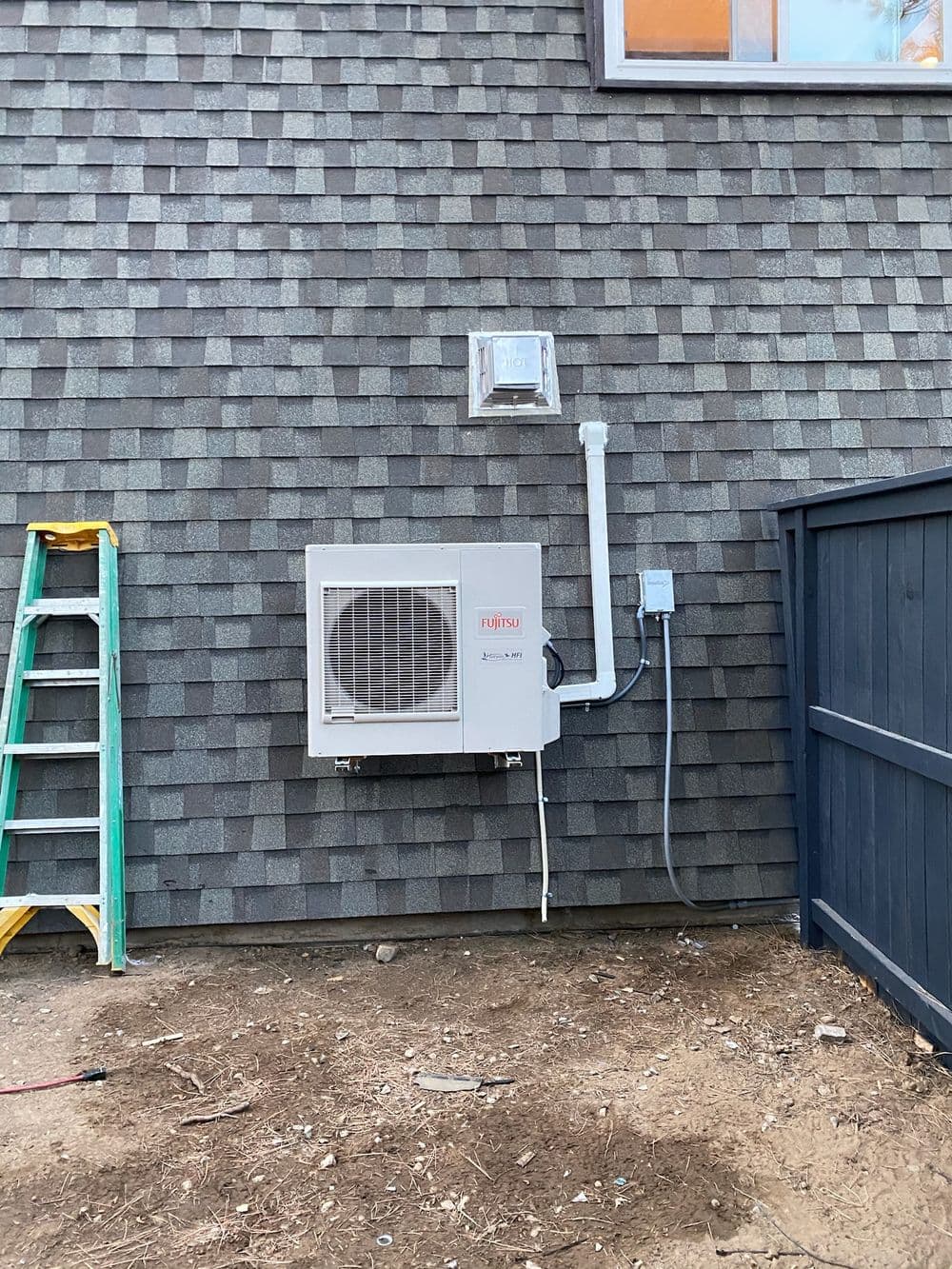 Air conditioning unit mounted on exterior wall of a house with ladder nearby and grey shingles.