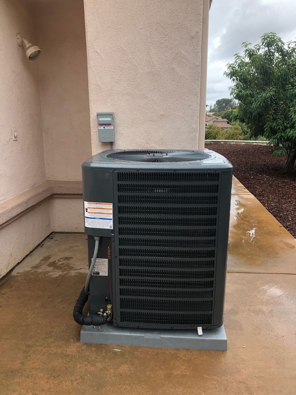 Air conditioning unit installed outside a building, surrounded by a concrete surface and greenery.