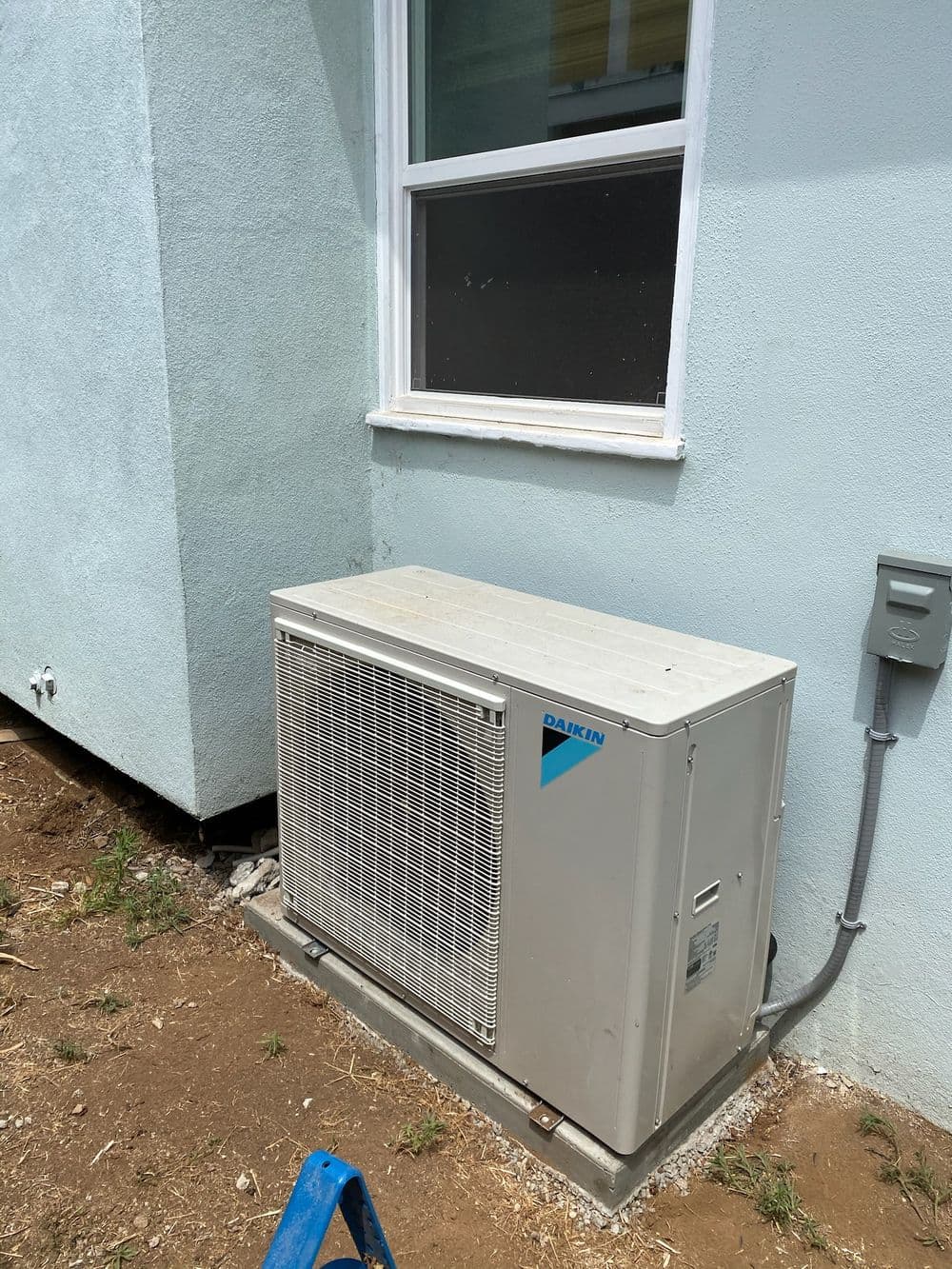 Daikin outdoor air conditioning unit installed beside a house wall with ladder in the foreground.