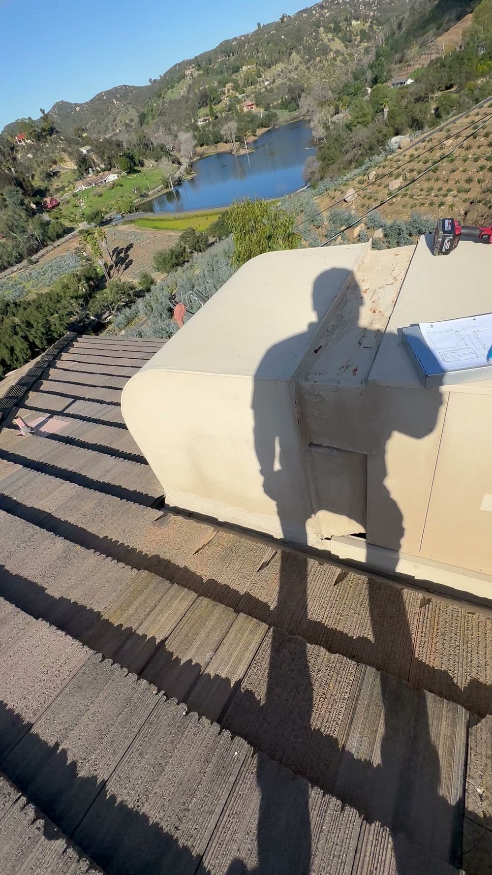 Person's shadow on a rooftop overlooking a lake and hillside landscape with vegetation.