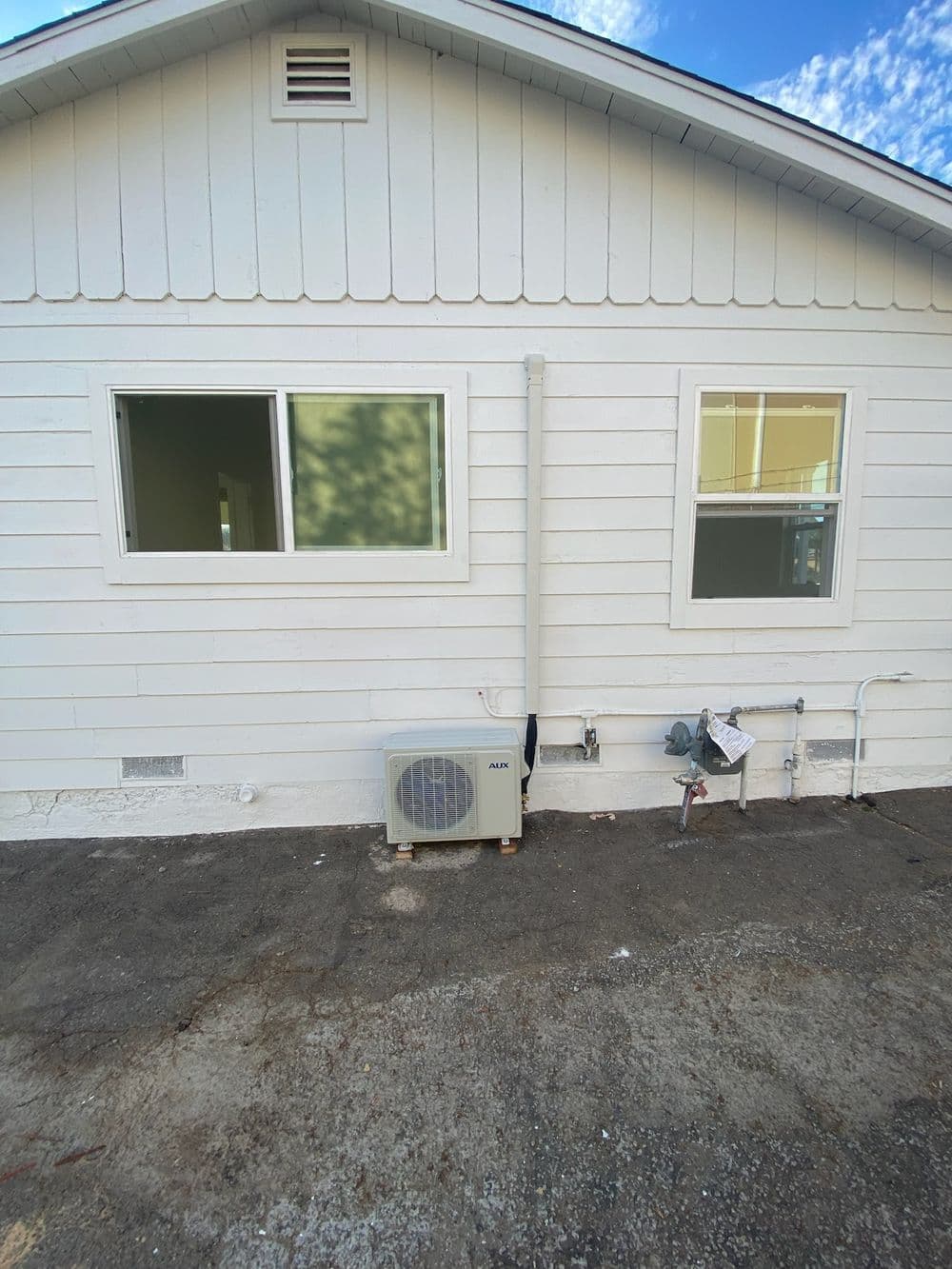 Air conditioning unit installed on the exterior wall of a white house with two windows.
