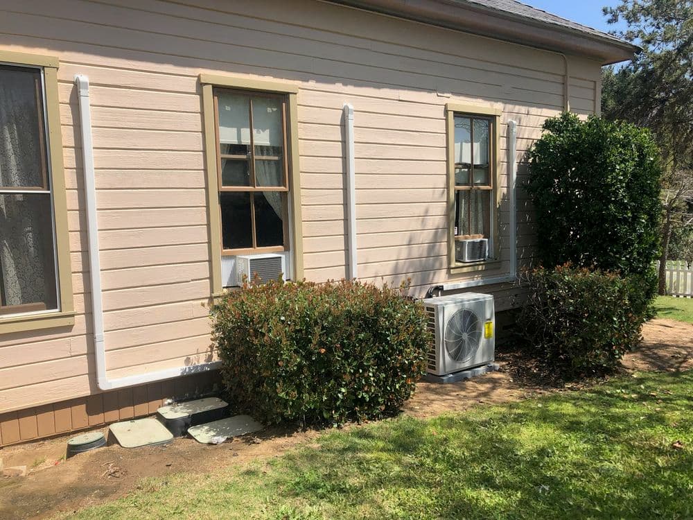 Air conditioning unit installed beside a light pink house with windows and green landscaping.