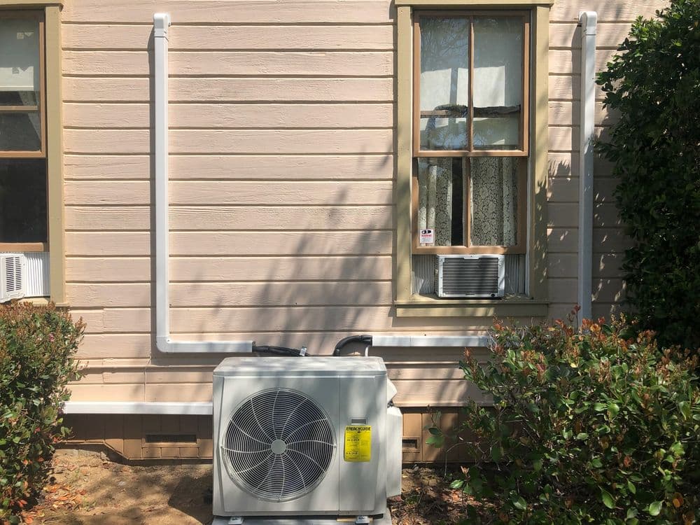 Air conditioning unit installed outside a house with white piping and window unit above.