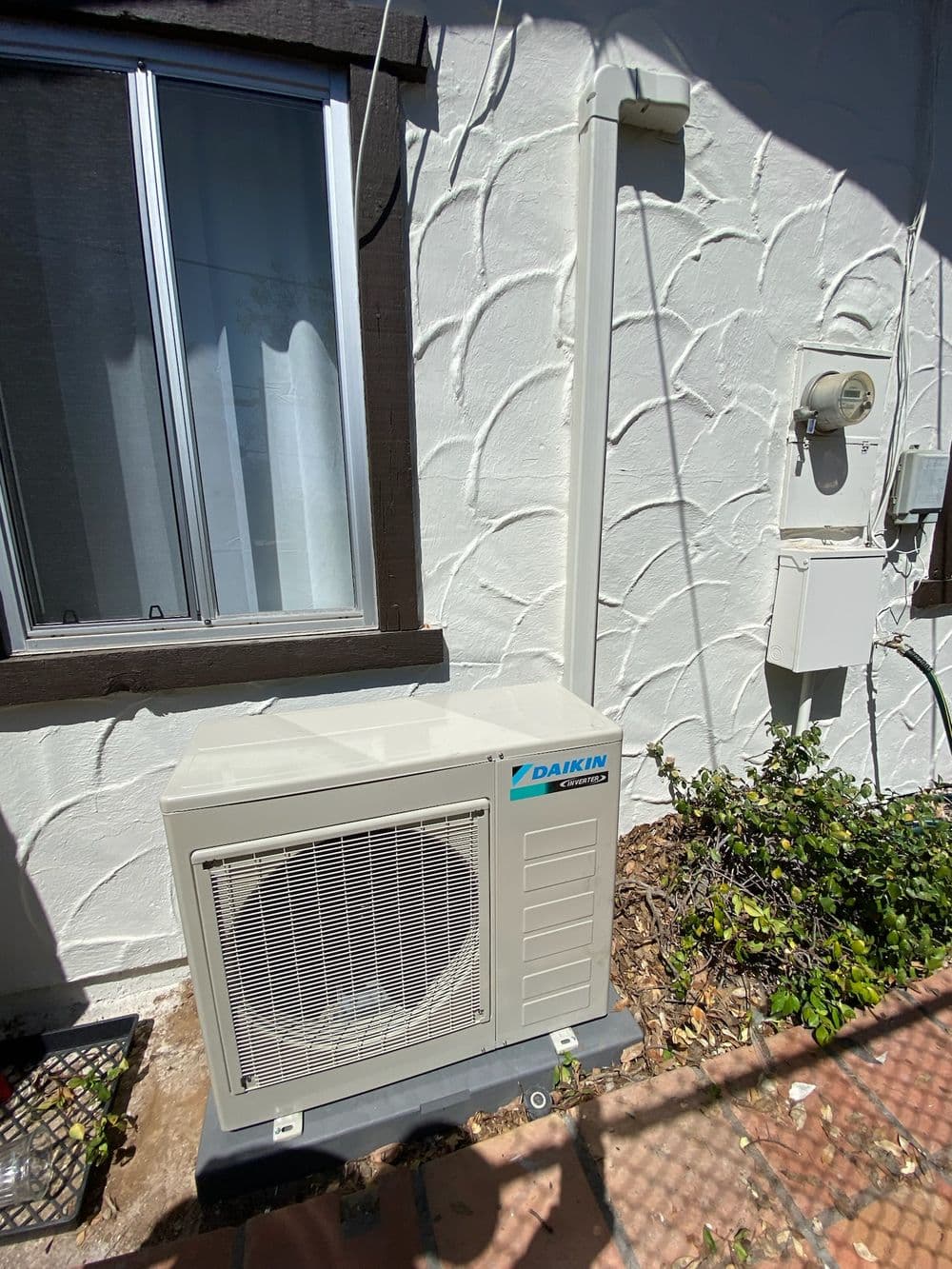 Daikin air conditioning unit installed on exterior wall with window and utility meter nearby.