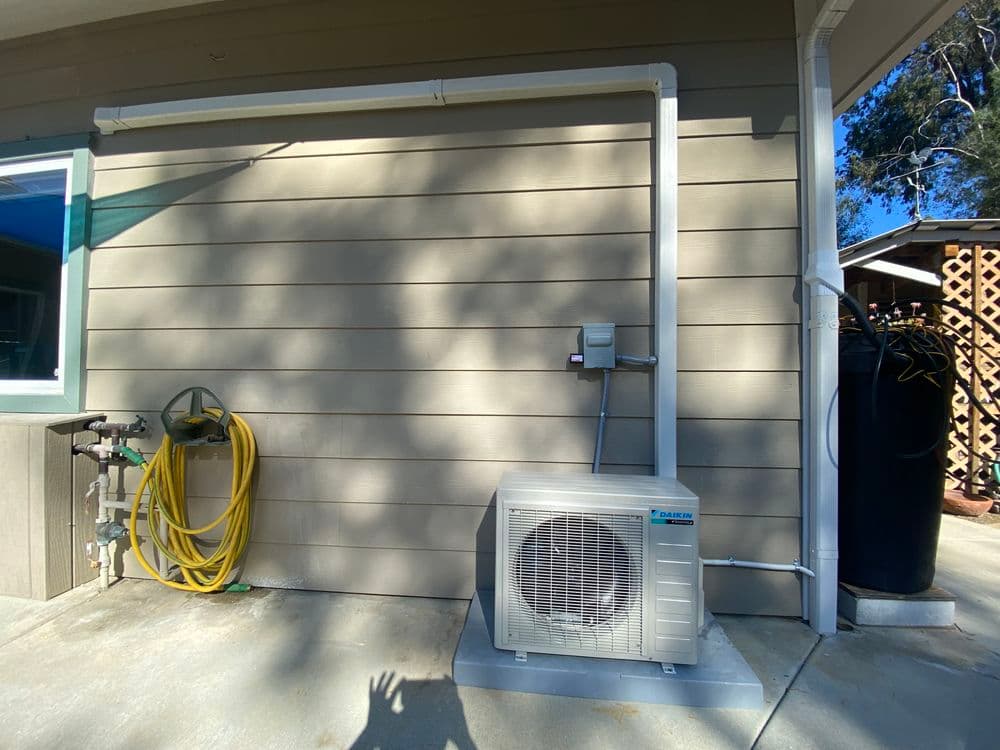 Air conditioning unit installed on exterior wall with hoses and utility connections nearby.