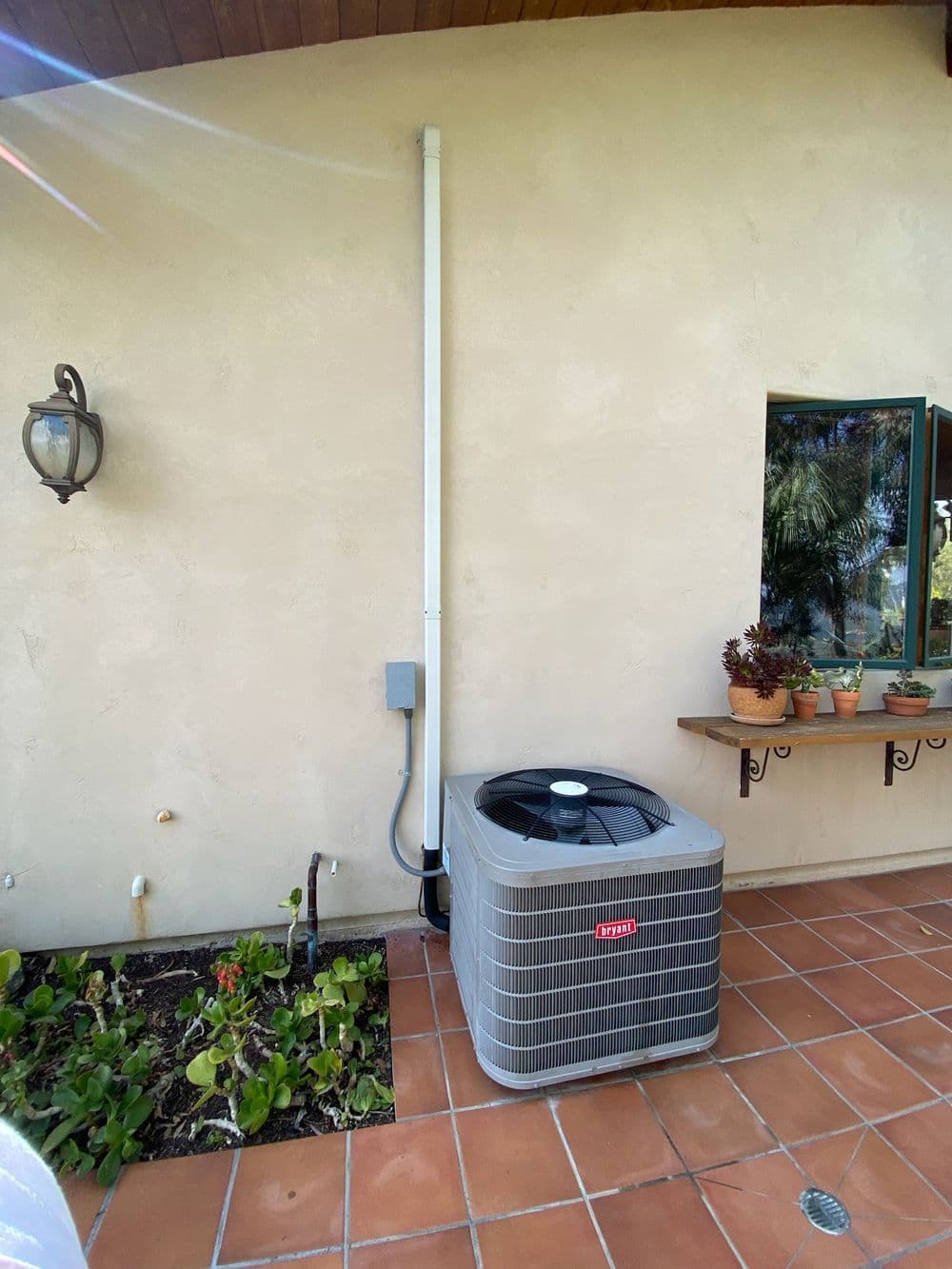 Air conditioning unit installed outside a house, surrounded by potted plants and tiles.