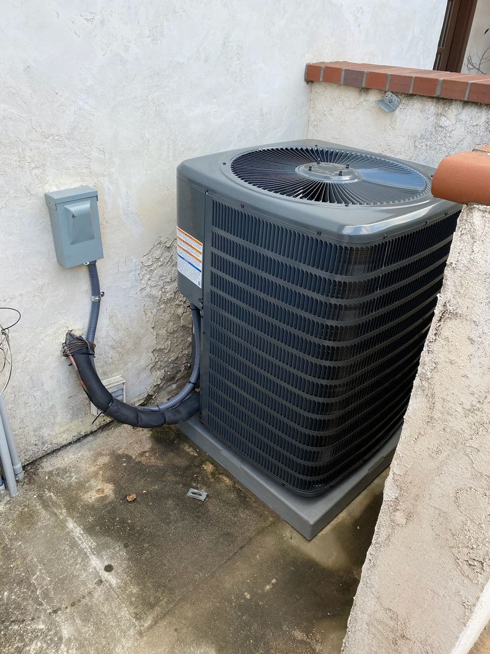 Residential air conditioning unit installed against a wall, with electrical wiring visible.