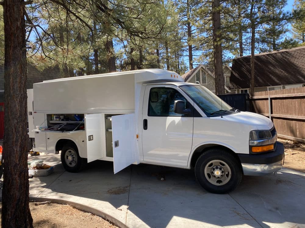 White service truck parked in wooded area, showcasing open side compartments for equipment.