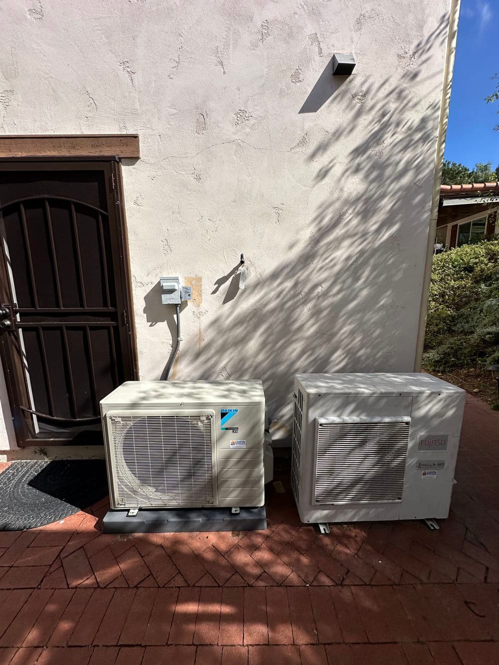 Air conditioning units installed outside a home, with a wall-mounted electrical panel nearby.