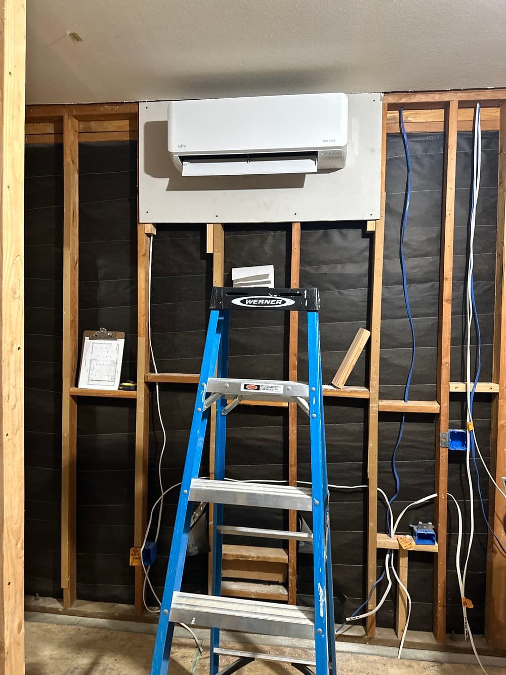 Ladder in front of wall-mounted air conditioner installation with exposed wiring.
