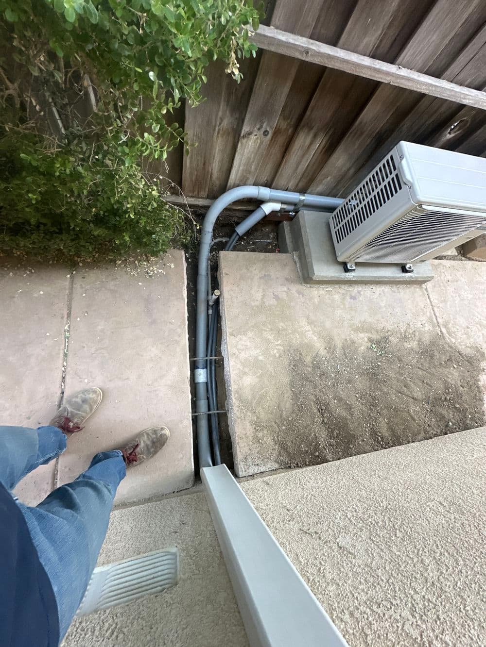 Person standing near a trench with pipes and an HVAC unit against a wooden fence.