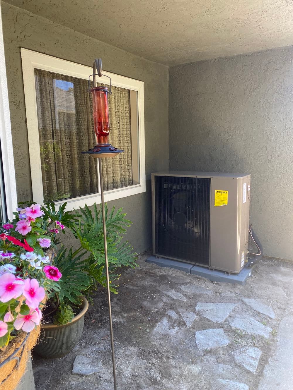 Air conditioning unit and hummingbird feeder in a cozy outdoor patio setting with flowers.