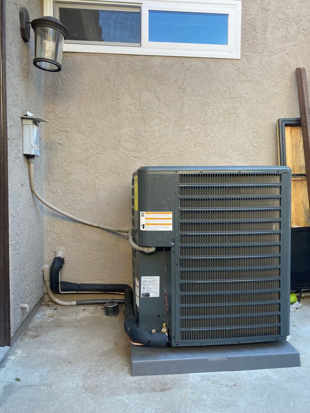 Air conditioning unit installed outside a home with adjacent power supply and light fixture.
