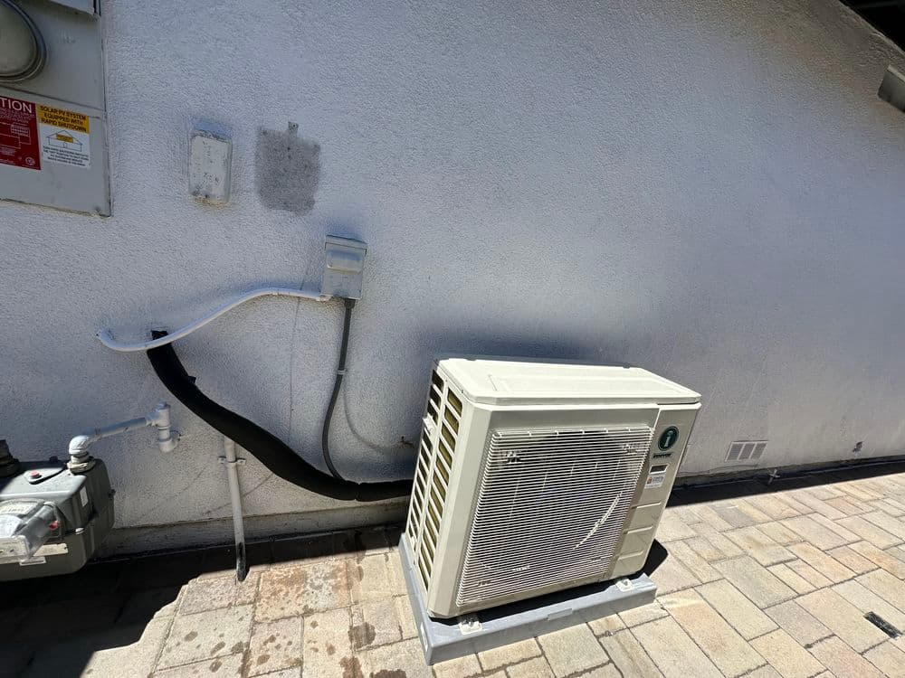 Air conditioning unit installed outside a building with white wall and brick flooring.