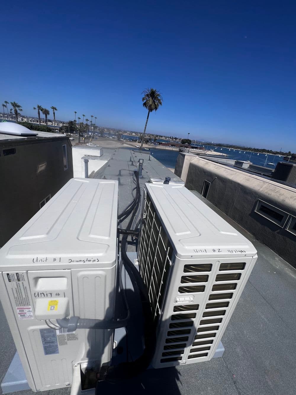 Rooftop view of air conditioning units with palm trees and waterfront in the background.